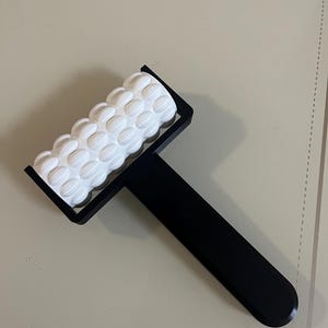 May include: A black and white massage roller with a textured white roller and a black handle. The roller has a series of rounded, raised bumps. The handle is long and rectangular.