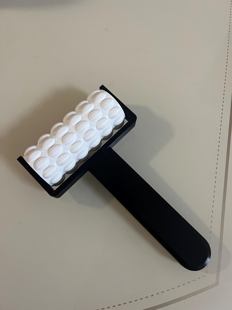 May include: A black and white massage roller with a textured white roller and a black handle. The roller has a series of rounded, raised bumps. The handle is long and rectangular.