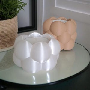 May include: Two bubble-shaped candle holders. One is white, the other is tan. The candle holders are sitting on a round, clear glass table. A potted plant is in the background.