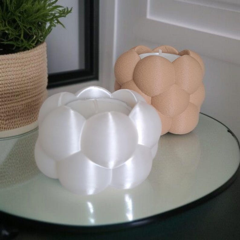 May include: Two bubble-shaped candle holders. One is white, the other is tan. The candle holders are sitting on a round, clear glass table. A potted plant is in the background.