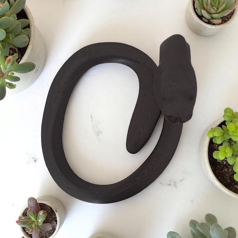 Puede incluir: Una escultura decorativa de serpiente negra mate. La escultura est&aacute; enrollada en forma circular con la cabeza levantada. Peque&ntilde;as suculentas en macetas rodean la escultura sobre una superficie de m&aacute;rmol blanco.