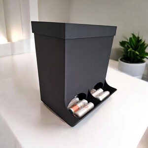 May include: A black, rectangular storage box with a slanted front, featuring two circular openings that hold several cylindrical items. The box is on a white surface, with a small plant in the background.