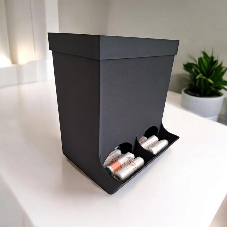 May include: A black, rectangular storage box with a slanted front, featuring two circular openings that hold several cylindrical items. The box is on a white surface, with a small plant in the background.