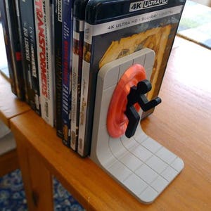 May include: A gray and orange portal-themed bookend supporting a stack of DVDs and Blu-ray discs. The bookend features a stylized figure emerging from an orange portal. The DVDs are on a wooden table.