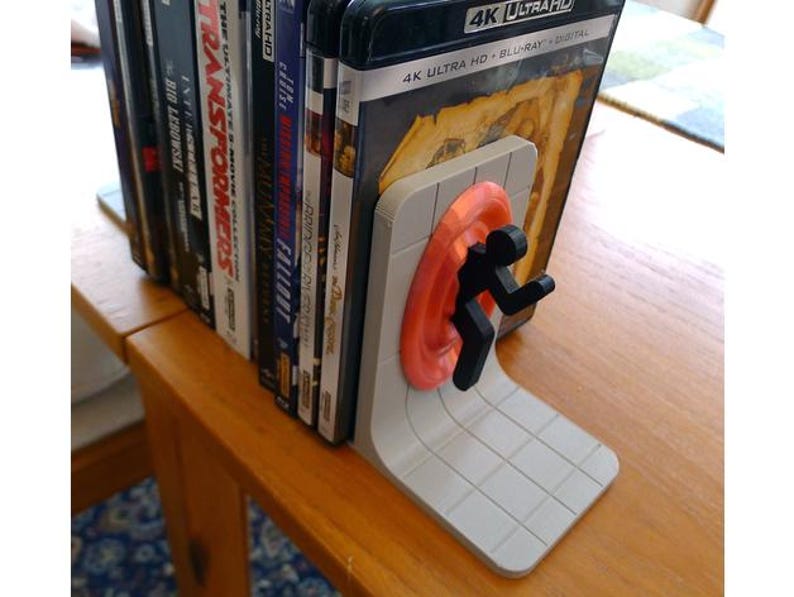 May include: A gray and orange portal-themed bookend supporting a stack of DVDs and Blu-ray discs. The bookend features a stylized figure emerging from an orange portal. The DVDs are on a wooden table.