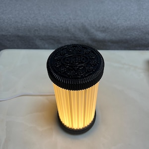 Pode incluir: Uma l&acirc;mpada em forma de biscoito Oreo preto e branco. A parte superior da l&acirc;mpada &eacute; um disco preto com o logotipo da Oreo. O corpo da l&acirc;mpada &eacute; um cilindro com pain&eacute;is verticais plissados ​​esbranqui&ccedil;ados que emitem um brilho quente. Um cabo branco se estende da base.