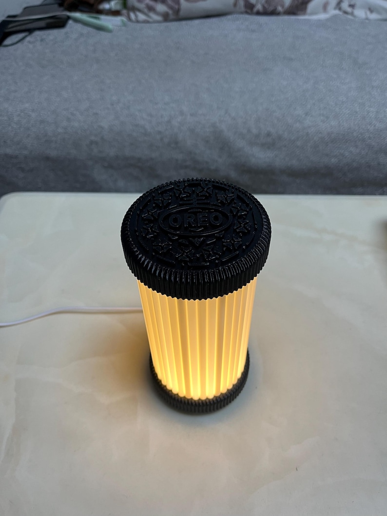 Pode incluir: Uma l&acirc;mpada em forma de biscoito Oreo preto e branco. A parte superior da l&acirc;mpada &eacute; um disco preto com o logotipo da Oreo. O corpo da l&acirc;mpada &eacute; um cilindro com pain&eacute;is verticais plissados ​​esbranqui&ccedil;ados que emitem um brilho quente. Um cabo branco se estende da base.
