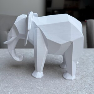 Op de afbeelding: Een witte, geometrische olifant sculptuur. De olifant is afgebeeld in een moderne, hoekige stijl met zichtbare facetten. De slagtanden en oren zijn duidelijk gedefinieerd. De sculptuur staat op een lichtgrijze ondergrond.