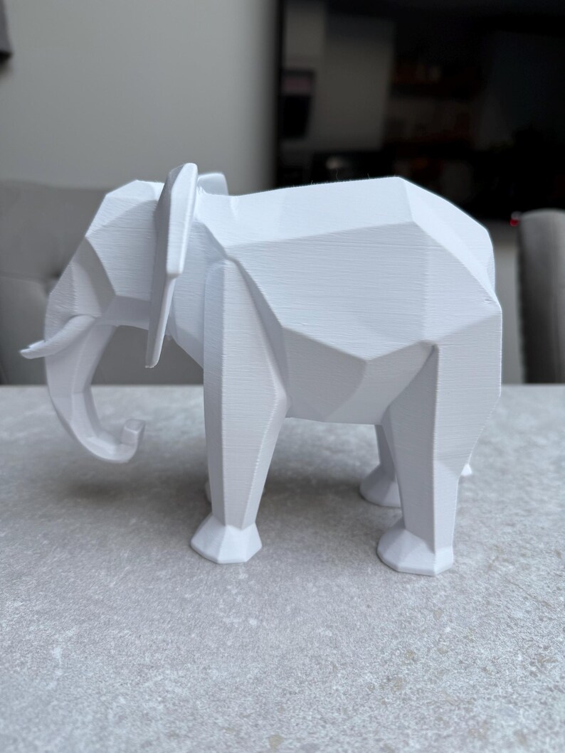 Op de afbeelding: Een witte, geometrische olifant sculptuur. De olifant is afgebeeld in een moderne, hoekige stijl met zichtbare facetten. De slagtanden en oren zijn duidelijk gedefinieerd. De sculptuur staat op een lichtgrijze ondergrond.