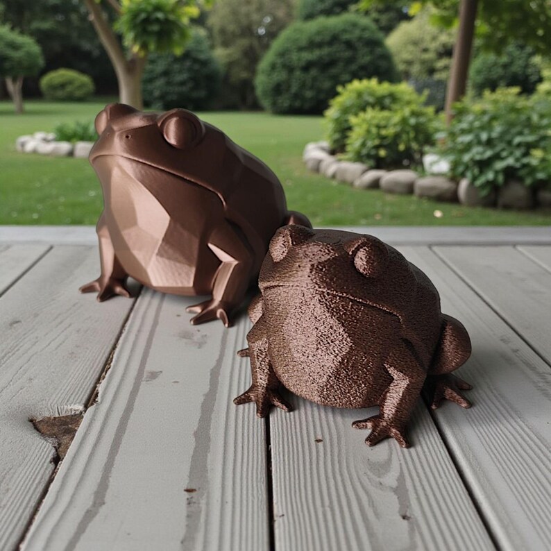 May include: Two bronze-colored frog sculptures. The larger frog has a geometric, faceted design, while the smaller one has a textured surface. Both frogs are positioned on a weathered wooden deck, with a green lawn and trees in the background.
