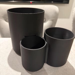 May include: A set of three black cylindrical containers of varying sizes. The largest is tall and straight, the middle one is shorter, and the third is angled. The containers are made of a matte black material.