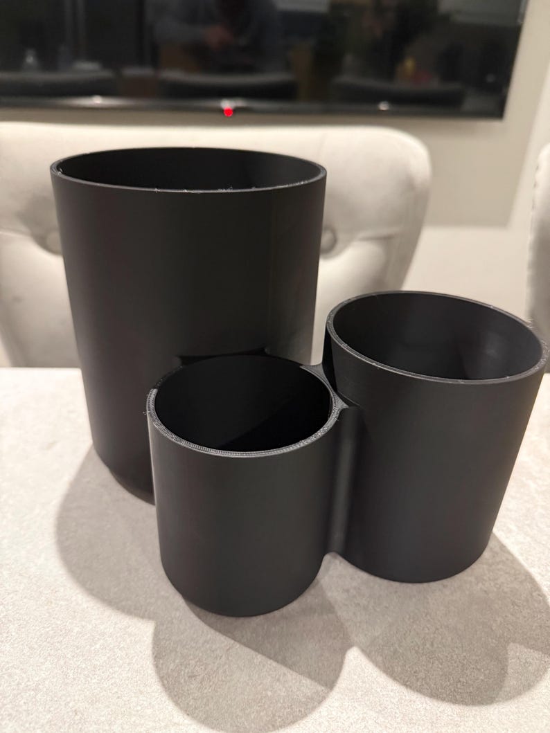 May include: A set of three black cylindrical containers of varying sizes. The largest is tall and straight, the middle one is shorter, and the third is angled. The containers are made of a matte black material.