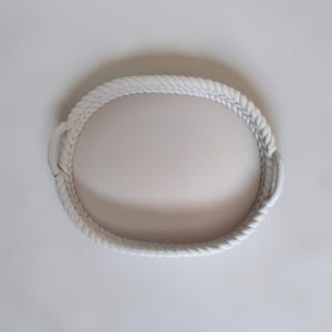 Puede incluir: Una bandeja ovalada blanca con un dise&ntilde;o de cuerda trenzada. La bandeja tiene dos asas peque&ntilde;as en lados opuestos. El patr&oacute;n de cuerda crea una superficie texturizada. La bandeja se encuentra sobre un fondo blanco liso.