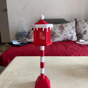Op de afbeelding: Een rood-witte decoratieve lantaarn met een vierkant kooiontwerp, bekroond met een witte bal en een sneeuwachtige rand. De lantaarn staat op een rood-witte gestreepte paal en een rode basis. Het item is waarschijnlijk voor vakantiedecoratie.