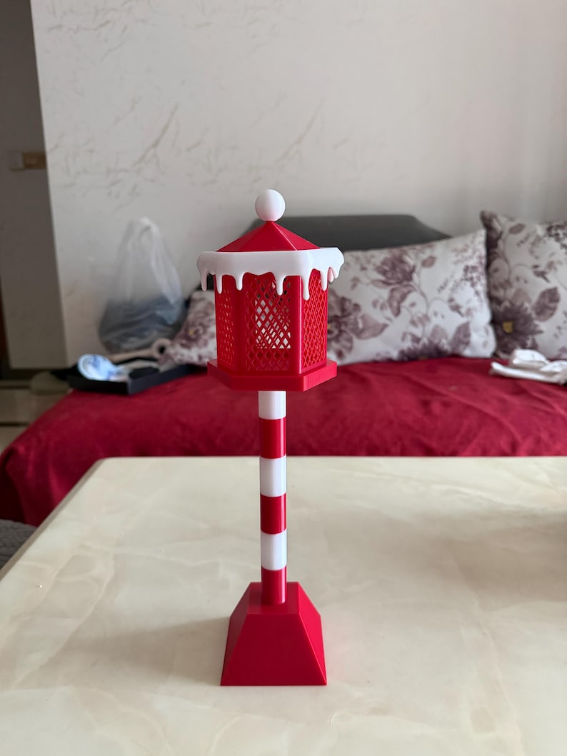 Op de afbeelding: Een rood-witte decoratieve lantaarn met een vierkant kooiontwerp, bekroond met een witte bal en een sneeuwachtige rand. De lantaarn staat op een rood-witte gestreepte paal en een rode basis. Het item is waarschijnlijk voor vakantiedecoratie.