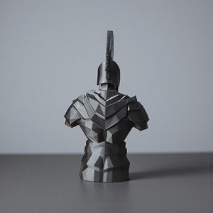 May include: A grey, geometric bust of a warrior, seen from the rear. The sculpture features a helmet with a tall crest and detailed armour. The design has a modern, angular aesthetic, with sharp lines and facets.