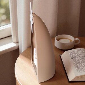May include: A beige and white decorative lamp with a star-shaped accent, on a wooden table. A cup of coffee and an open book are also on the table, near a window with a curtain.