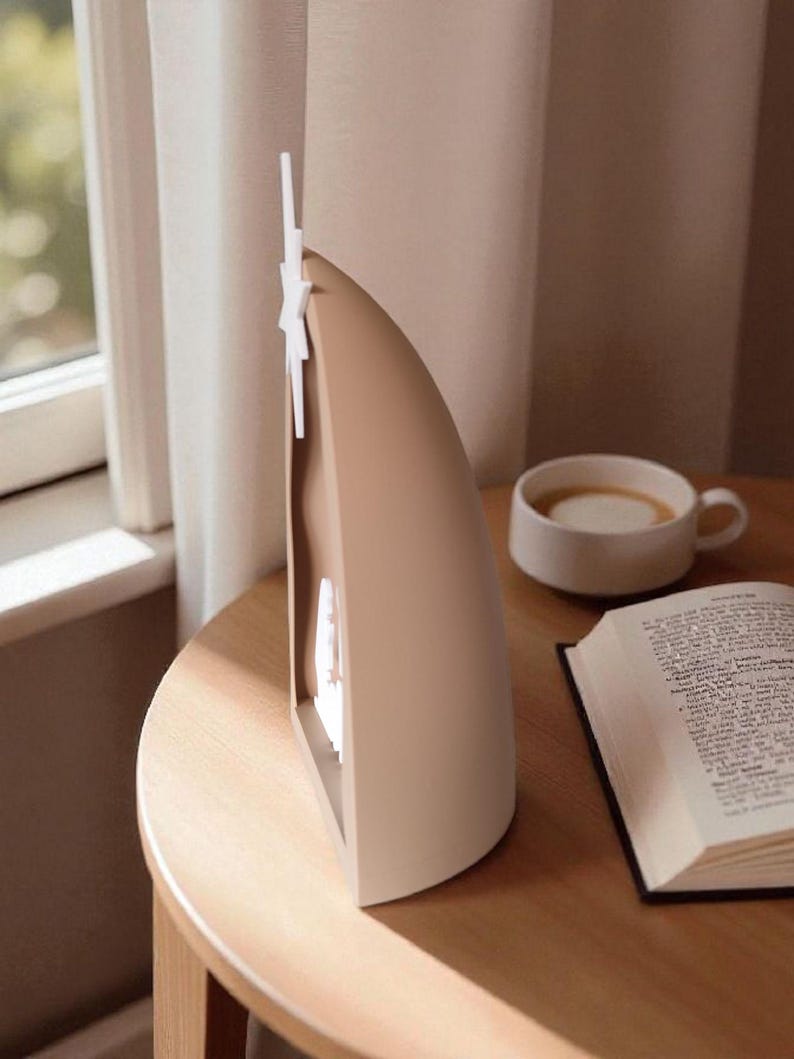May include: A beige and white decorative lamp with a star-shaped accent, on a wooden table. A cup of coffee and an open book are also on the table, near a window with a curtain.