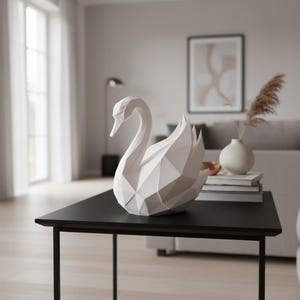 May include: A white geometric swan sculpture with a detailed, angular design. The swan is positioned on a black table, showcasing its elegant form. The sculpture is a decorative home accent, adding a modern touch to the interior.