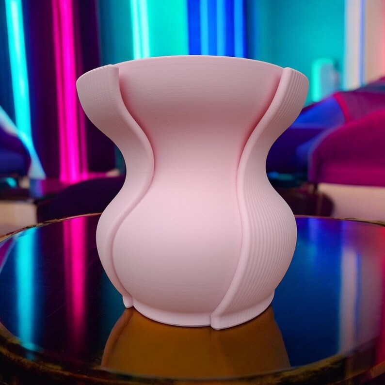 Puede incluir: Un jarr&oacute;n rosa claro con un dise&ntilde;o curvo &uacute;nico. El jarr&oacute;n tiene una abertura ancha y una base redondeada. Los lados del jarr&oacute;n tienen un patr&oacute;n texturizado y acanalado. El jarr&oacute;n est&aacute; sobre una superficie reflectante.