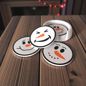 May include: Set of four snowman-themed coasters with black rims. Each coaster features a different snowman face design with orange carrot noses and black eyes or mouths. A white coaster holder is also included.