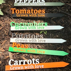 May include: A collection of colorful garden markers, each labeled with the names of vegetables: Peppers, Tomatoes, Cucumbers, Potatoes, Peas, and Carrots. The text "Grown with love" is printed below each vegetable name. The markers are in a garden bed.