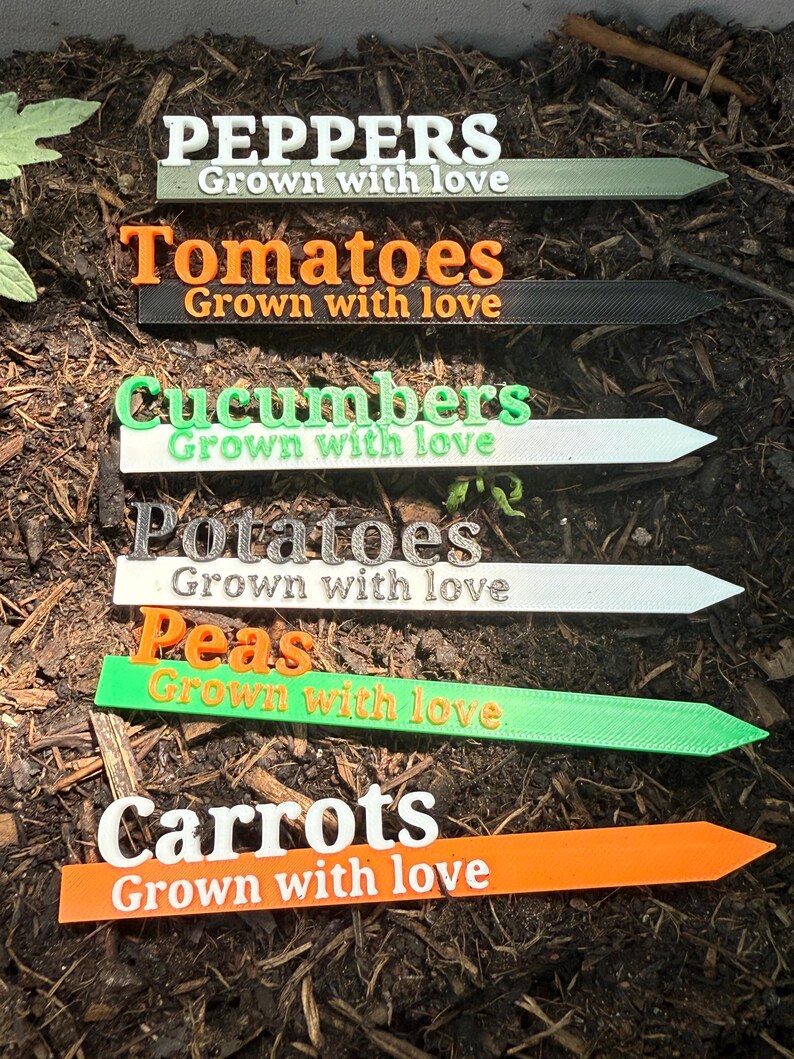 May include: A collection of colorful garden markers, each labeled with the names of vegetables: Peppers, Tomatoes, Cucumbers, Potatoes, Peas, and Carrots. The text "Grown with love" is printed below each vegetable name. The markers are in a garden bed.