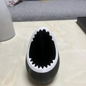 May include: A black and white shark-shaped container. The container has a black body with a white, jagged edge around the opening, resembling a shark's mouth. The interior is black.
