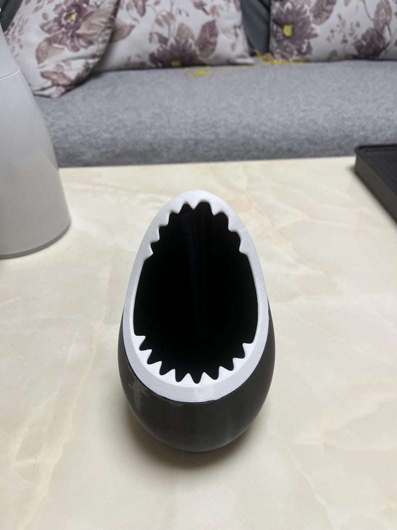 May include: A black and white shark-shaped container. The container has a black body with a white, jagged edge around the opening, resembling a shark's mouth. The interior is black.