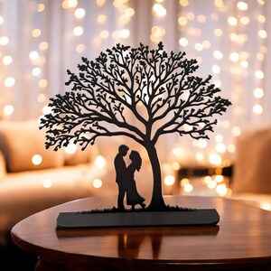May include: A black metal sculpture of a tree with a couple embracing beneath its branches. The tree has intricate, detailed branches and sits on a rectangular base. The background features soft, blurred lights.