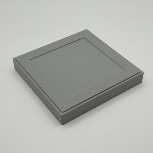 Pu&ograve; includere: Una scatola quadrata grigia con una parte superiore incassata e strutturata e un bordo rialzato. La scatola &egrave; di un colore grigio opaco e sembra essere fatta di un materiale rigido. Il design &egrave; semplice e moderno.