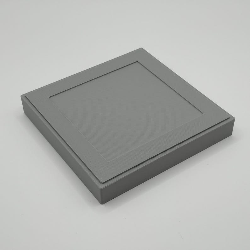 Pu&ograve; includere: Una scatola quadrata grigia con una parte superiore incassata e strutturata e un bordo rialzato. La scatola &egrave; di un colore grigio opaco e sembra essere fatta di un materiale rigido. Il design &egrave; semplice e moderno.
