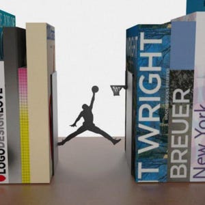 May include: A set of decorative bookends featuring a silhouette of a basketball player mid-dunk. The bookends are positioned on either side of the silhouette, with colorful book spines visible. The bookends are designed to hold books upright.