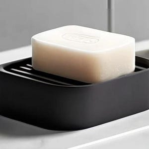 May include: A black soap dish with a rectangular shape, holding a bar of white soap. The soap dish has a slotted design to allow for drainage. The soap is a light beige color and has a slightly curved top.