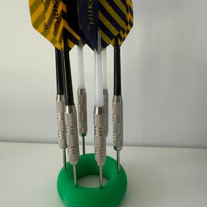 May include: A collection of darts with yellow and black striped flights, marked "NUKE", are held in a green circular stand. The darts have silver metal barrels and black shafts. The stand is on a white surface.