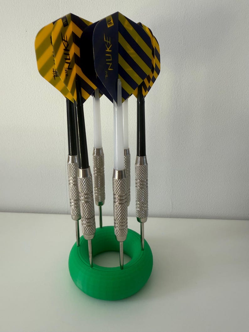May include: A collection of darts with yellow and black striped flights, marked "NUKE", are held in a green circular stand. The darts have silver metal barrels and black shafts. The stand is on a white surface.