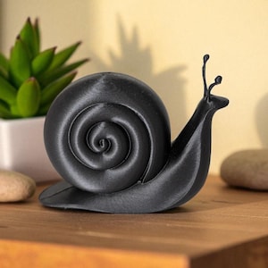 May include: A black snail figurine with a spiraled shell and two antennae. The snail is made of a matte material and sits on a wooden surface. A small succulent plant in a white pot is in the background.
