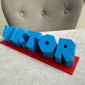 Op de afbeelding: Een blauwe 3D-naam, "VIKTOR", wordt weergegeven op een rode rechthoekige basis. De letters zijn blokkerig en staan rechtop. De achtergrond toont een grijs oppervlak en een grijs gestoffeerde stoel.