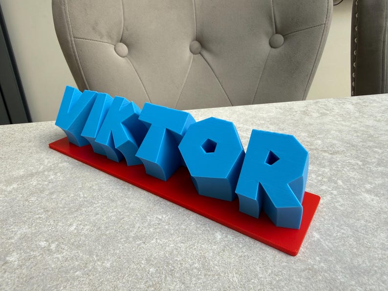 Op de afbeelding: Een blauwe 3D-naam, "VIKTOR", wordt weergegeven op een rode rechthoekige basis. De letters zijn blokkerig en staan rechtop. De achtergrond toont een grijs oppervlak en een grijs gestoffeerde stoel.