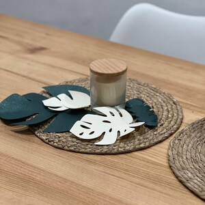 May include: A decorative table setting featuring a woven placemat, a candle in a glass jar with a wooden lid, and several leaf-shaped cutouts in white and dark green. The arrangement is on a wooden table.