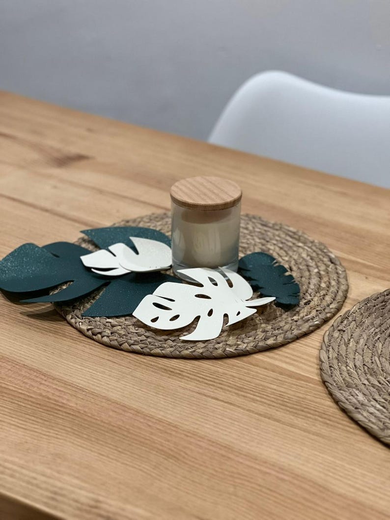 May include: A decorative table setting featuring a woven placemat, a candle in a glass jar with a wooden lid, and several leaf-shaped cutouts in white and dark green. The arrangement is on a wooden table.