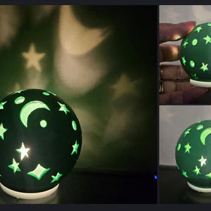May include: A dark green sphere-shaped lamp with cut-out star, moon, and dot designs. The lamp sits on a white base and projects star and moon shapes onto the surrounding surface. The lamp is a decorative night light.