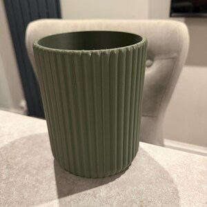 May include: A sage green, ribbed plant pot. The cylindrical pot has vertical ridges and sits on a light-colored surface. The pot is empty and ready for planting. The background includes a chair and a wall.