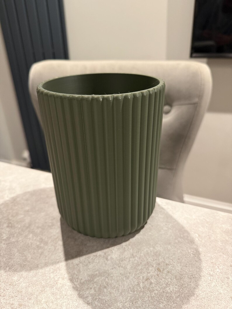 May include: A sage green, ribbed plant pot. The cylindrical pot has vertical ridges and sits on a light-colored surface. The pot is empty and ready for planting. The background includes a chair and a wall.