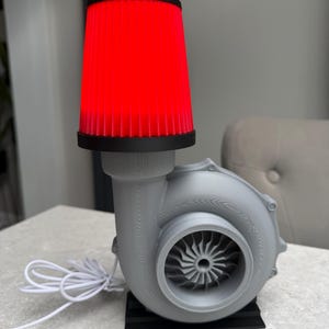 May include: A gray, 3D-printed lamp shaped like a turbocharger, with a bright red filter as the lampshade. The lamp sits on a black base and has a white power cord.