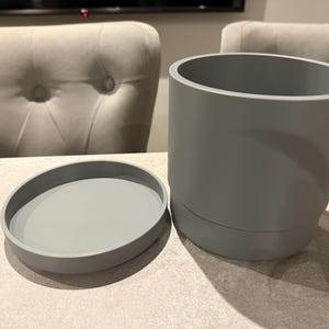 May include: A gray cylindrical planter with a matching saucer. The planter is approximately 6 inches tall and 6 inches in diameter. The saucer is about 1 inch tall and 7 inches in diameter. The items are made of a smooth, solid material.