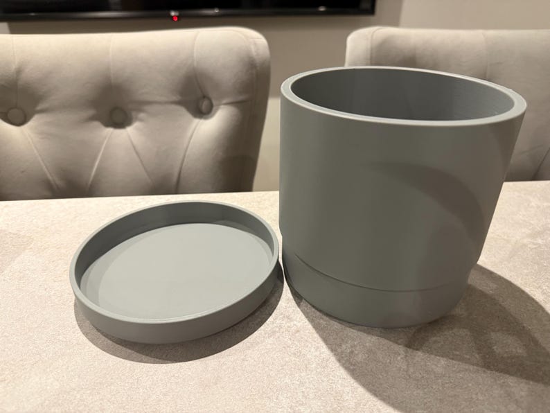 May include: A gray cylindrical planter with a matching saucer. The planter is approximately 6 inches tall and 6 inches in diameter. The saucer is about 1 inch tall and 7 inches in diameter. The items are made of a smooth, solid material.