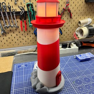 May include: A red and white lighthouse model with a light inside. The lighthouse has a red top and a white and red striped body. It sits on a gray base with a rock-like design. The lighthouse is approximately 10 inches tall.