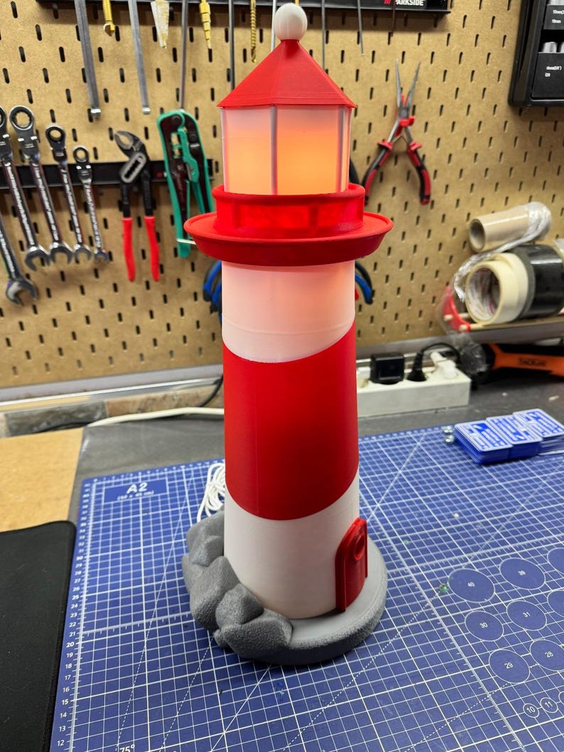 May include: A red and white lighthouse model with a light inside. The lighthouse has a red top and a white and red striped body. It sits on a gray base with a rock-like design. The lighthouse is approximately 10 inches tall.