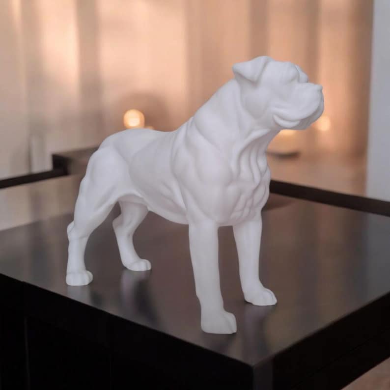May include: A white, stylized dog sculpture in a standing pose. The dog has a muscular build and a focused expression. The sculpture is placed on a dark, reflective surface, with a blurred background.